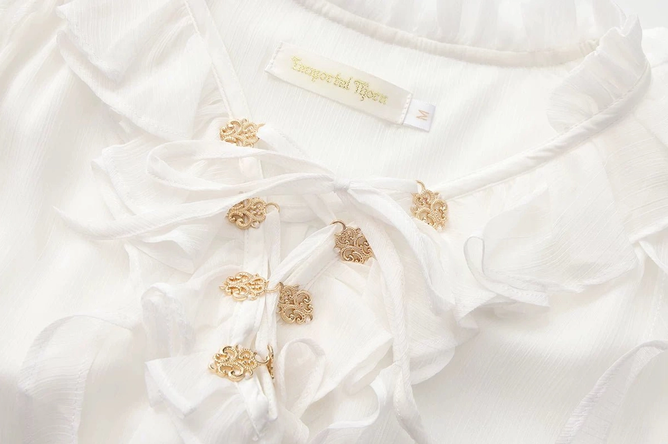 Close-up macro detail of white ouji lolita blouse ruffled bow front neckline trim with vintage gold decorative buttons and inner brand label on collar, top-down detail shot