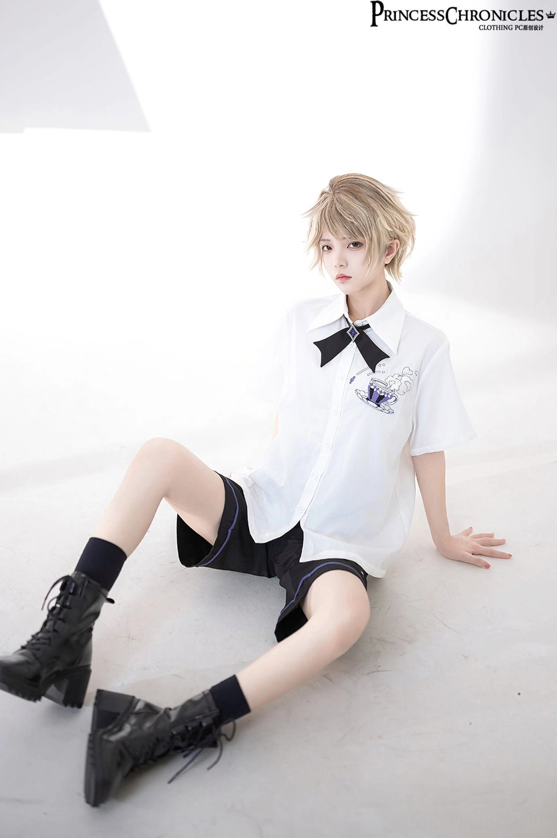 Androgynous model in ouji lolita outfit seated angle view showing embroidered short-sleeve shirt with bow tie and black combat boots socks