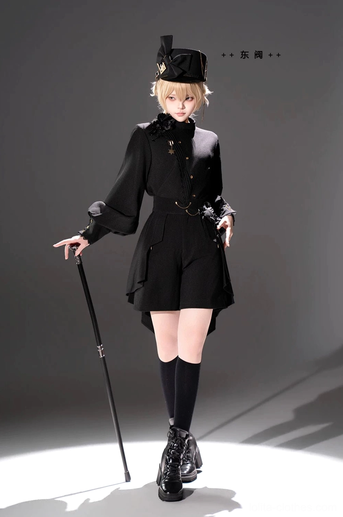 Ouji Lolita black uniform set front three-quarter view with blonde model in military top hat holding walking cane wearing black combat boots and structured black fabric