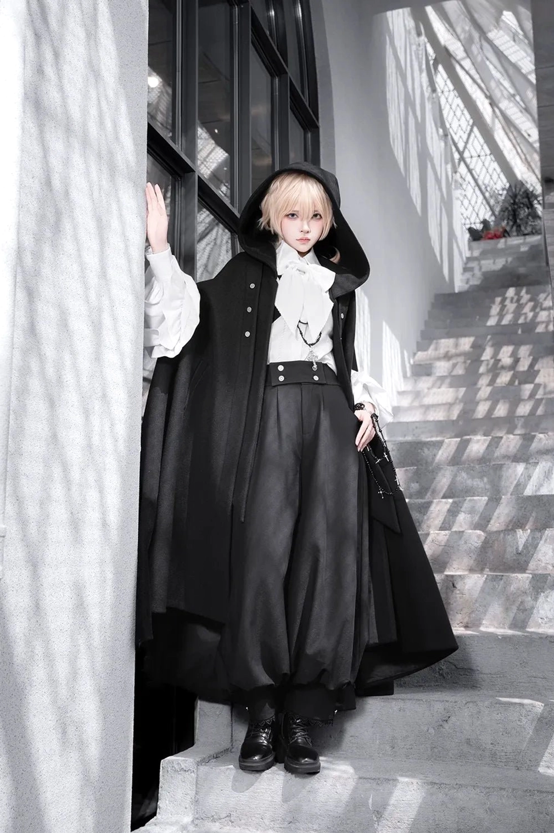Ouji lolita ensemble three-quarter side view model wearing hooded cape with waist belt and black leather shoes at indoor staircase