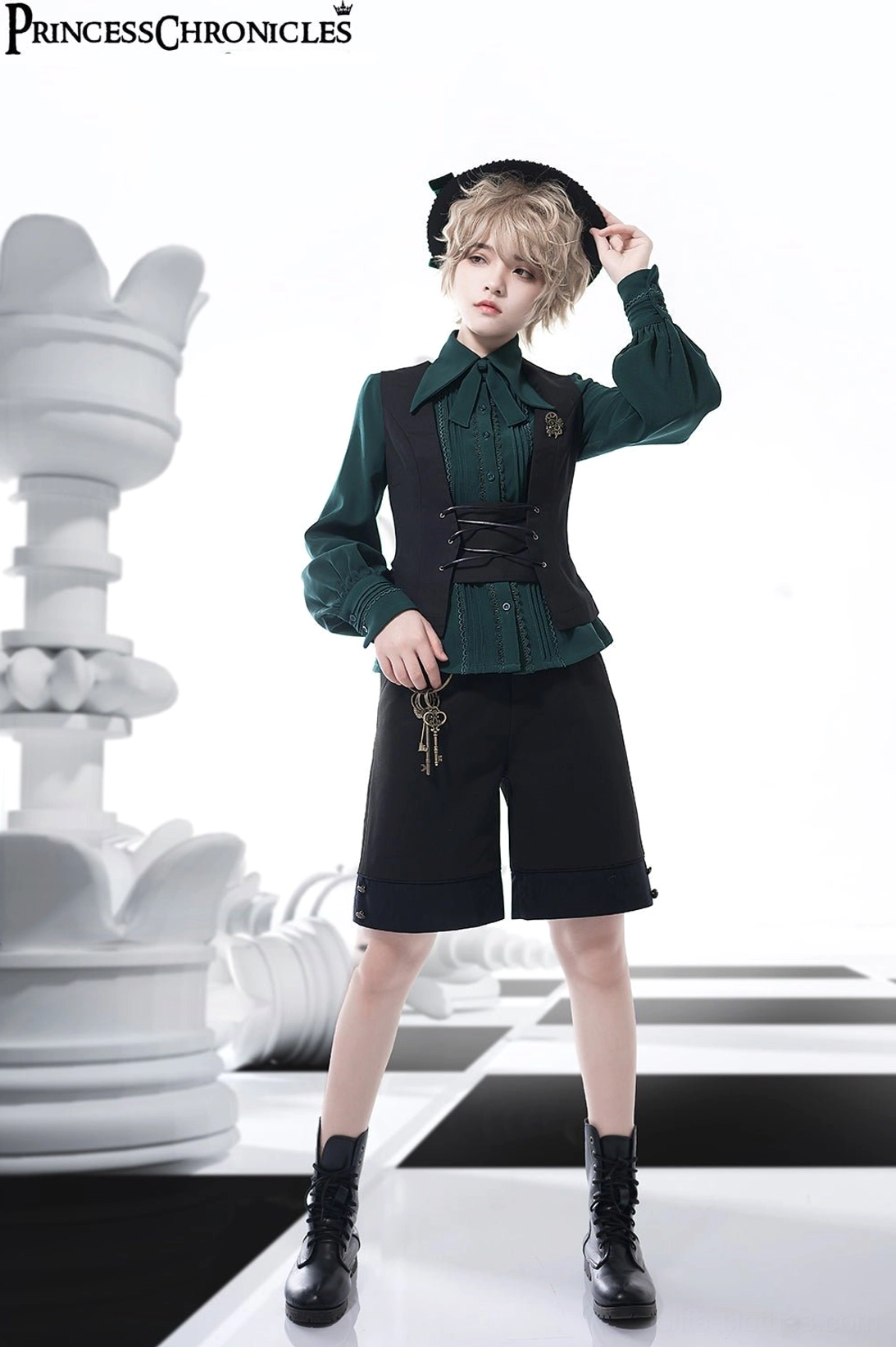 Full front standing view of androgynous model in black Ouji Lolita waistcoat with lace-up front over ruffled green blouse and chain decorated shorts