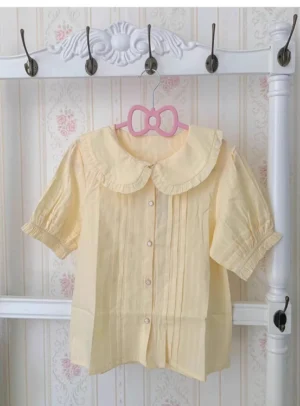 Pale yellow lolita cotton blouse short sleeve with ruffled collar and cuffs, front flat lay showing button closure and pleated front panels on vintage floral background
