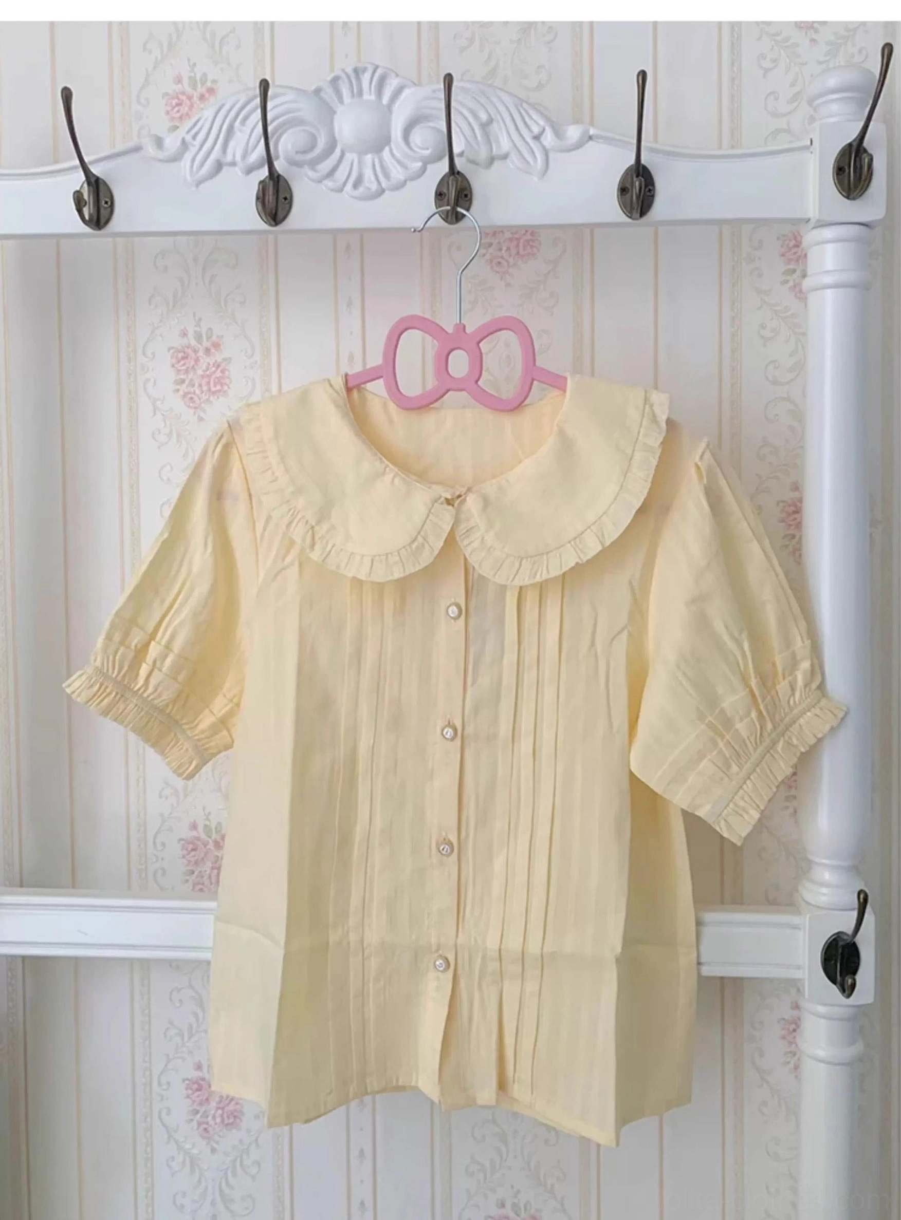 Pale yellow lolita cotton blouse short sleeve with ruffled collar and cuffs, front flat lay showing button closure and pleated front panels on vintage floral background