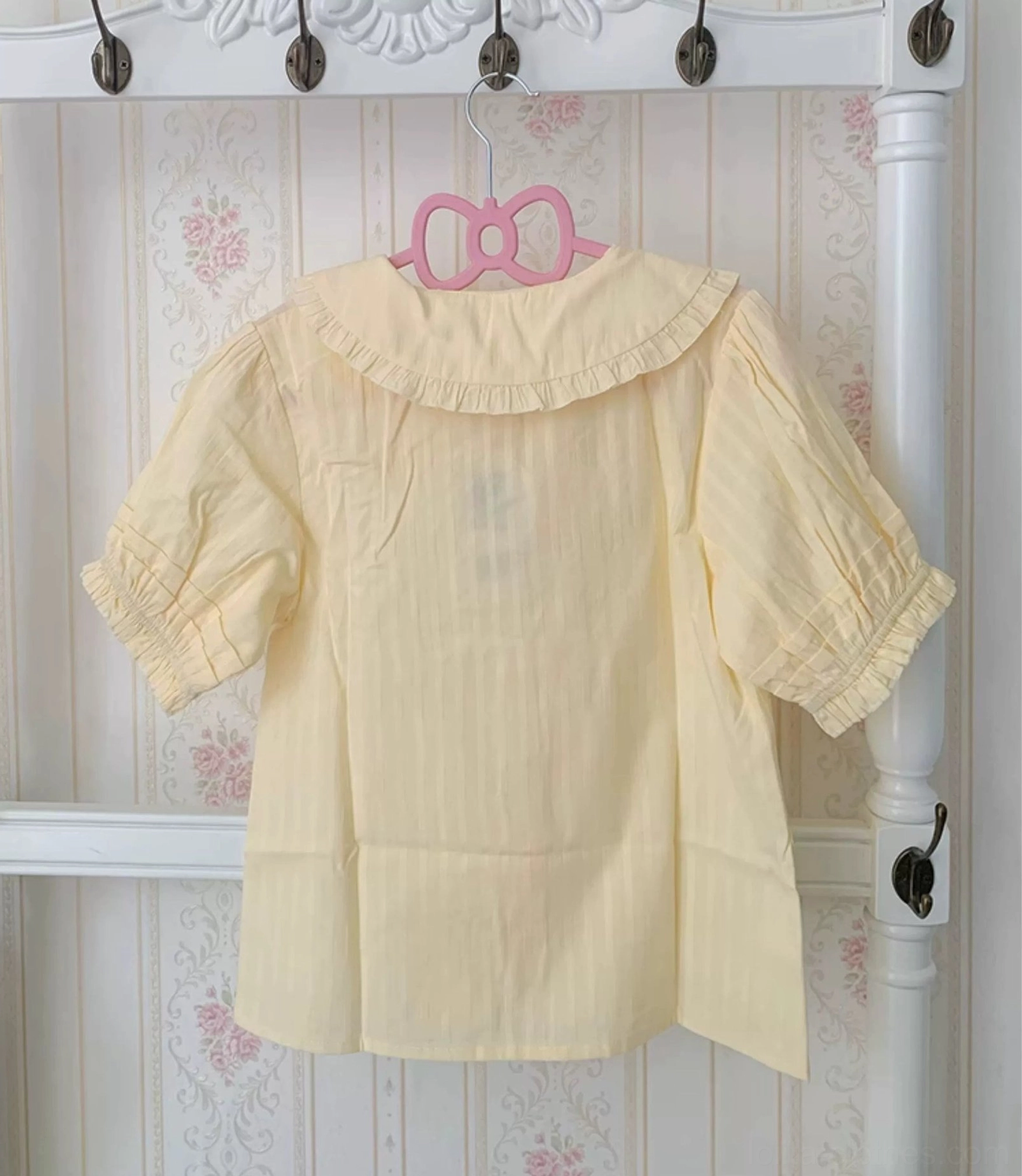 Pale yellow lolita blouse back view showing simple unadorned design, ruffled puff sleeves, and relaxed silhouette on hanger with vintage floral wallpaper backdrop