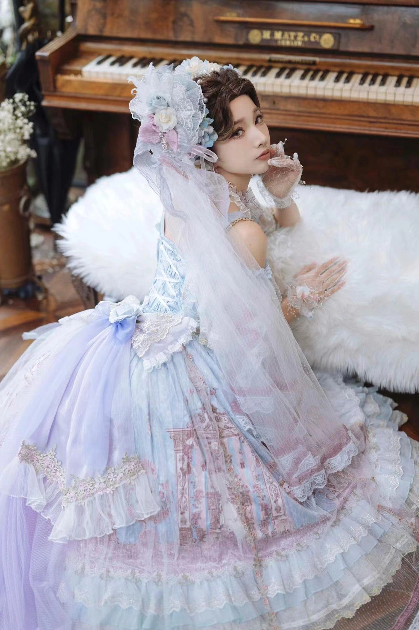 Pastel blue wedding Lolita JSK dress back view with large waist bow decoration and layered ruffled train skirt, model with floral veil headpiece on white fur sofa