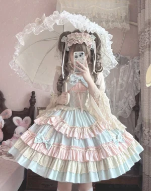 Front full-body view of pastel candy tiered ruffle sweet lolita jumper skirt with large chest bow and layered lace-trimmed skirt, worn by lolita girl in pink vintage bedroom with lace parasol and matching hair headband