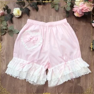 Pastel pink cotton lolita lace bloomers featuring heart-shaped lace motif and coordinating pink satin bow, multi-tiered ruffled lace hem with matching pink ribbon ties, romantic sweet lolita design, flat lay overhead shot on wooden surface with rose flower styling