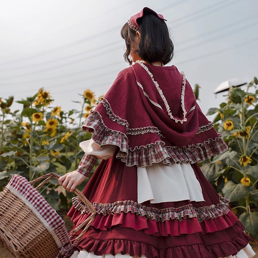 Full-body back view of model wearing hooded cape and pastoral Lolita OP dress in sunflower flower field, carrying checkered picnic basket, showcasing exposed ruffled trim details and wine red cloak construction