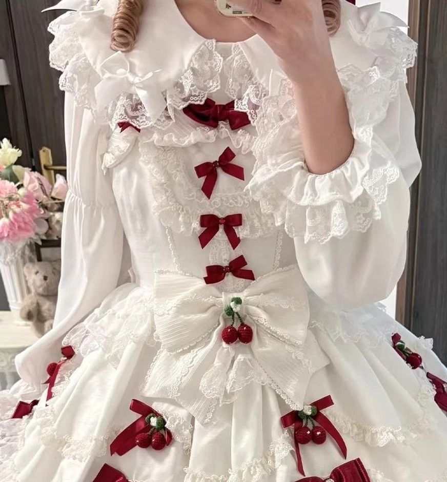 Over-the-shoulder mirror selfie of model wearing white Peter Pan collar sweet Lolita blouse upper half with front red bow lacing and large front bow with cherry ornaments