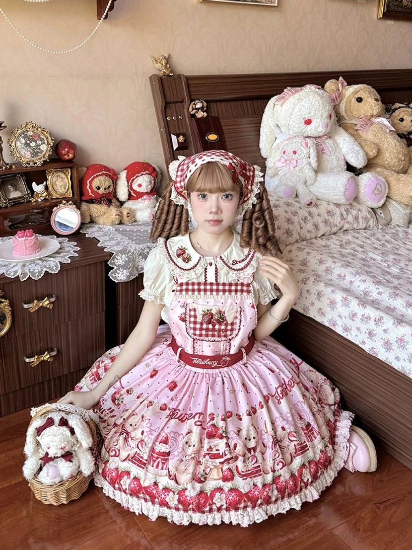 Pink-beige colorway lolita salopette dress with all-over strawberry and bear print, lace ruffled hem, gingham chest panel in vintage bedroom setting with plush toys