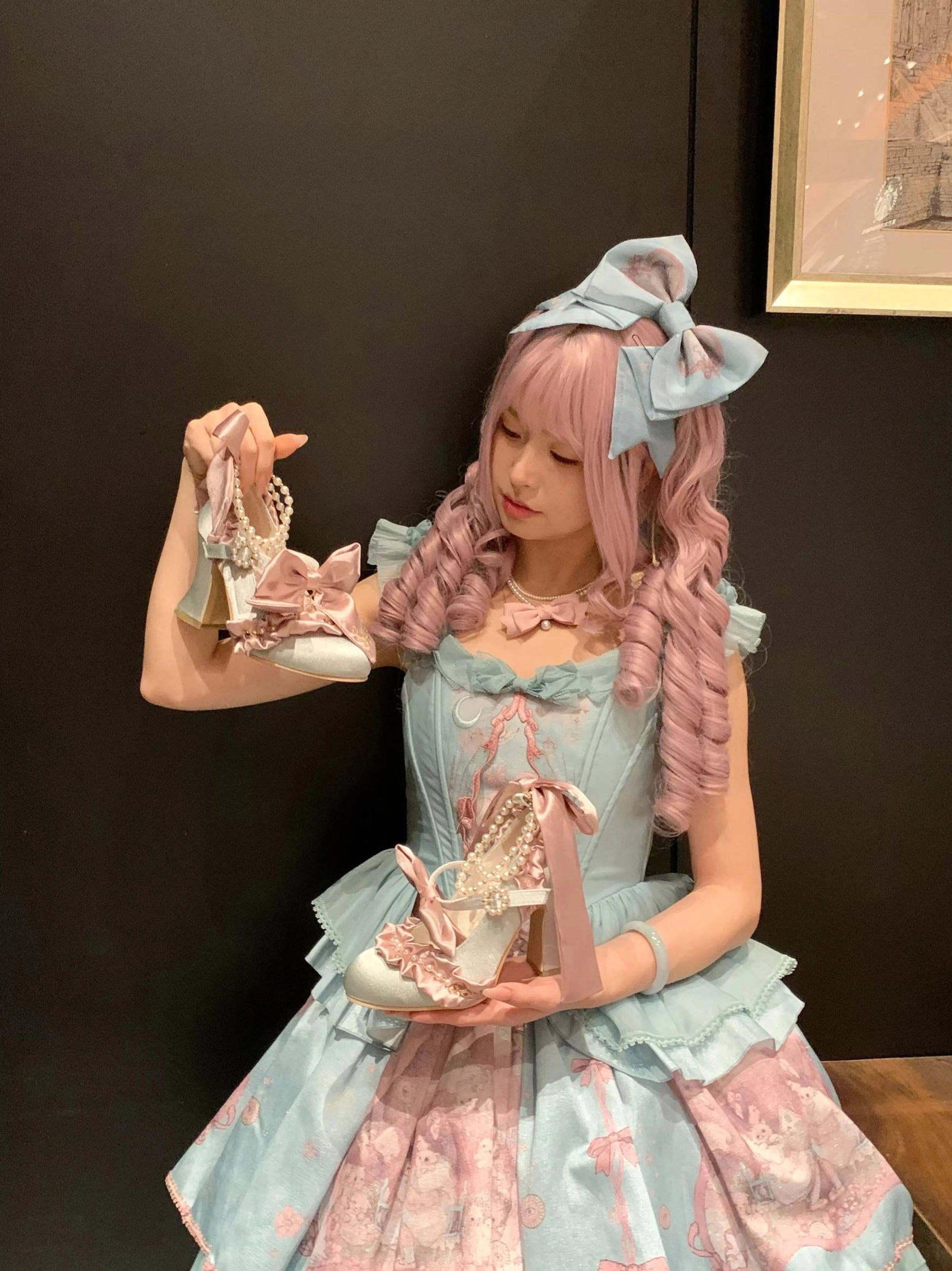 Pink-blue Classic Lolita block heel shoes with pearl ankle strap and detachable satin bow, model holding shoes in hands, styled with blue-pink Lolita dress and pink curly wig with bow headband