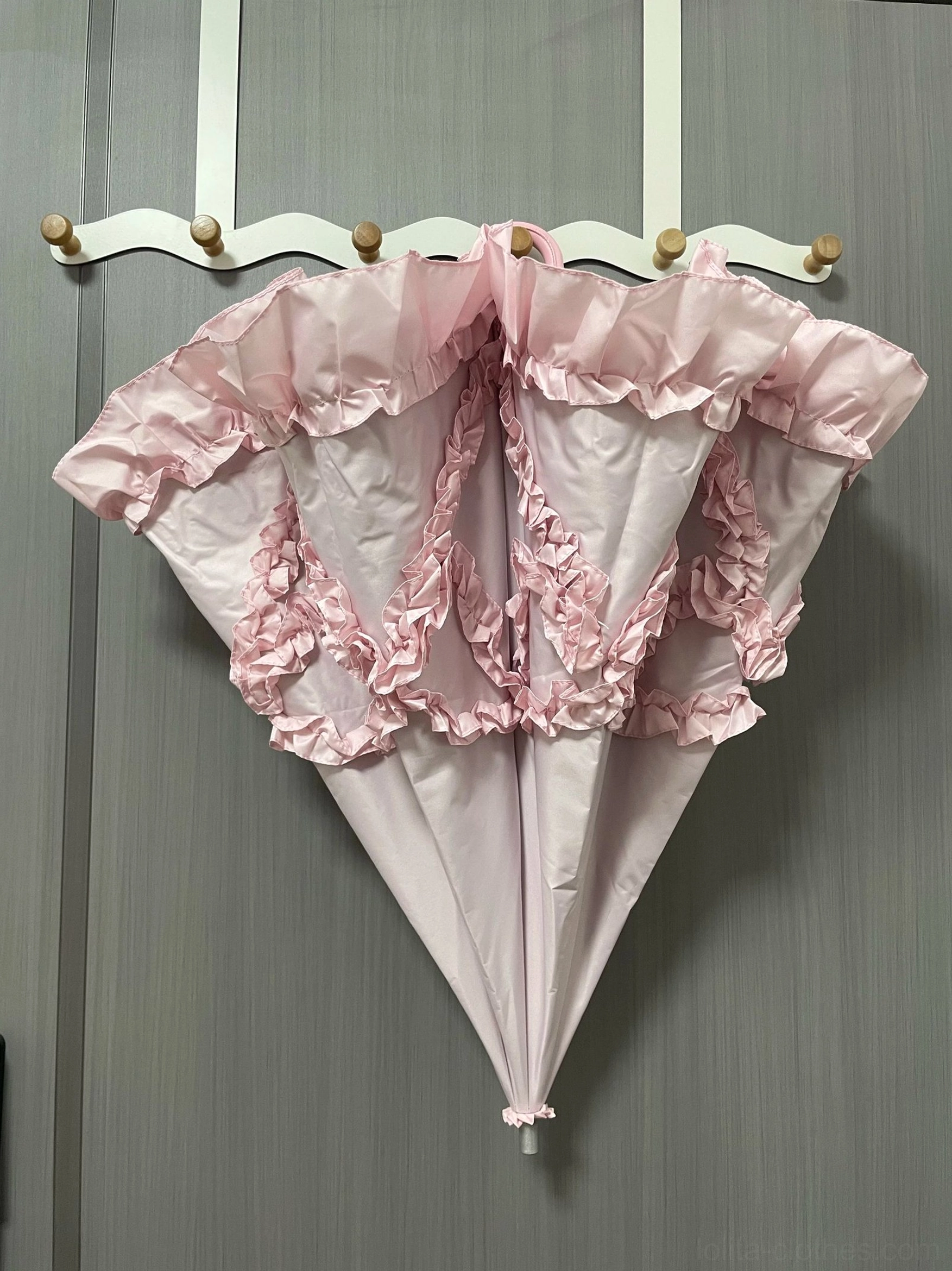 Pink bow-shaped ruffled Lolita parasol with gradient fabric and decorative 3D frill details displayed hanging on wall hooks, indoor home interior setting
