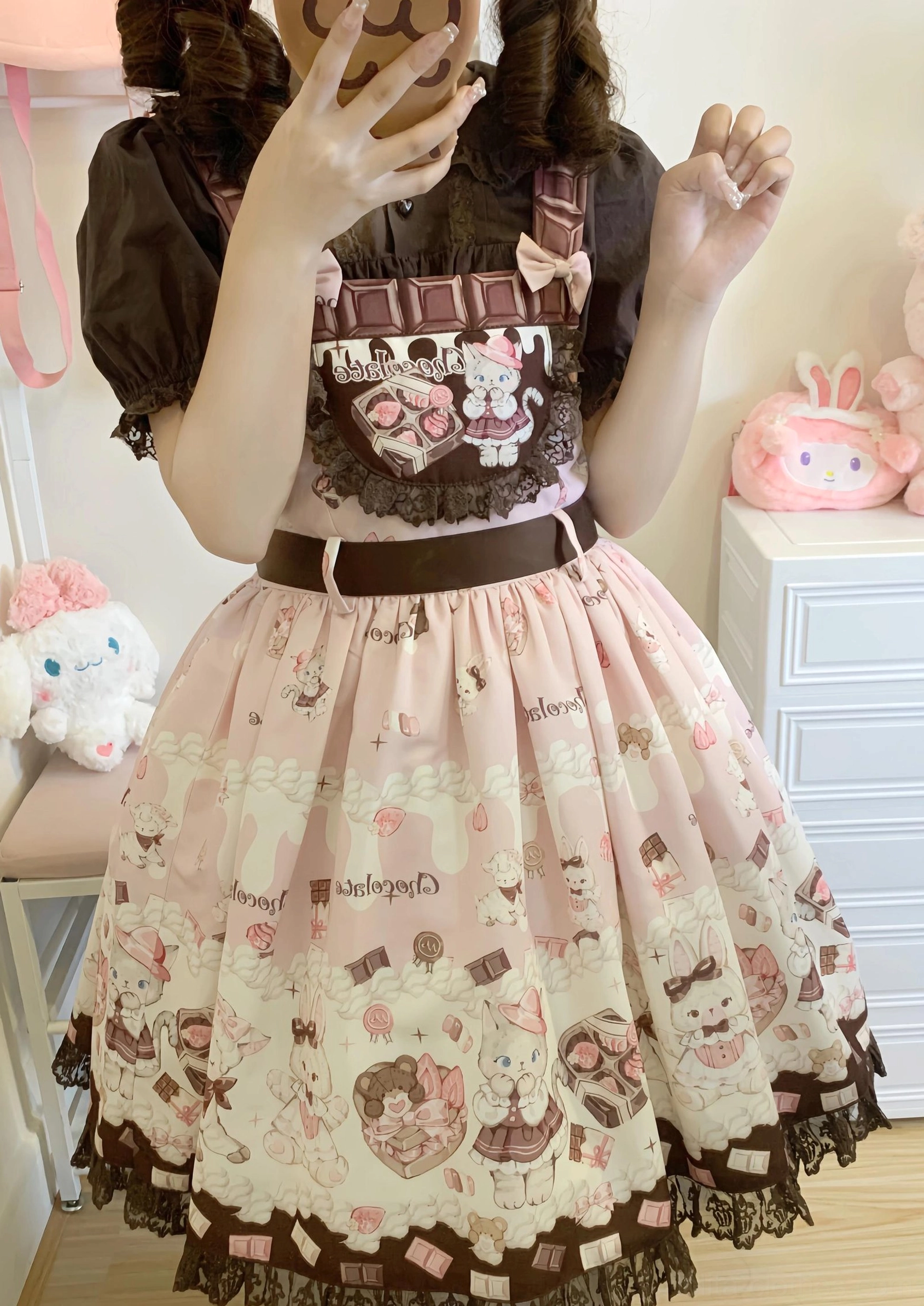 Model wearing pink and brown cute cat print Lolita salopette dress with puff sleeve inner blouse, waist bow belt, and pleated lace hem skirt, half-body front gesture pose in cozy bedroom setting
