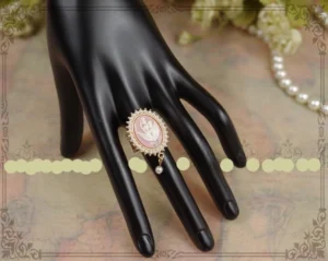 Pink cameo Lolita pearl ring with dangling pearl accent worn on black mannequin hand, displayed on vintage tabletop with roses and pearl necklace, ornate gold filigree frame setting