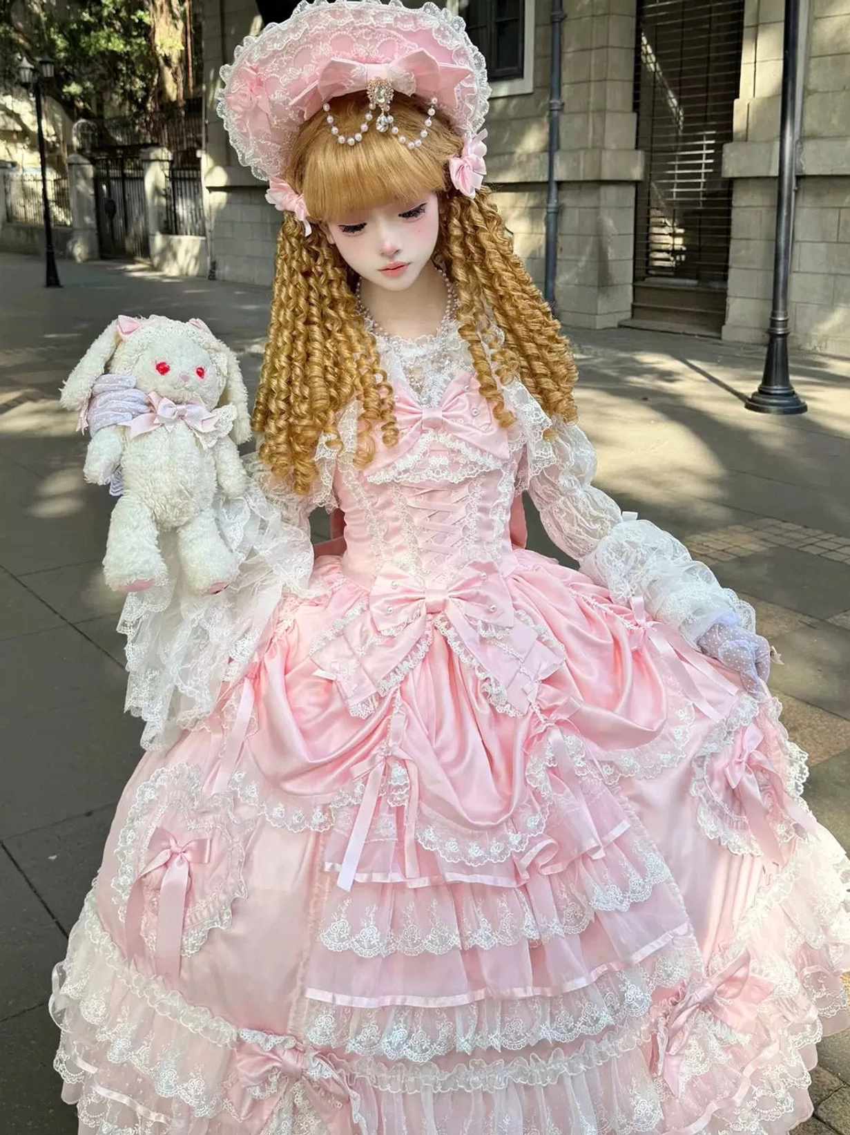 Pink sweet wedding Lolita JSK dress three-quarter front view with layered lace skirt, waist bow decorations, satin bodice, worn by blonde curly twin tails model with pink bonnet and white lace gloves in outdoor street setting