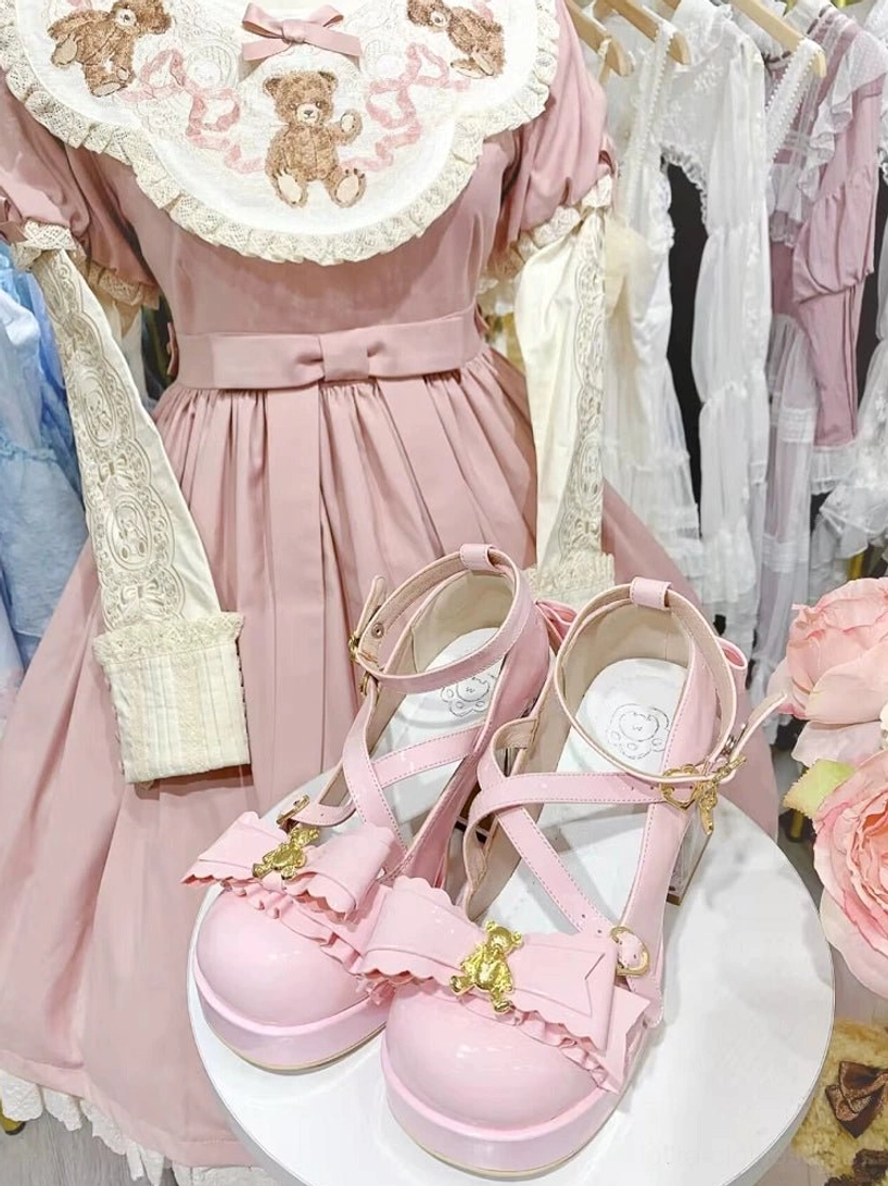 Pink sweet lolita platform heels on white display stand, front close-up view, pink teddy bear embroidery lolita dress backdrop, crisscross straps, gold bear charm, ruffled bow detail, clear block heel