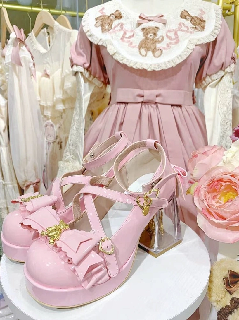 Pink sweet lolita platform heels three-quarter side shot on white display stand, pink lolita dress and rose flower decoration background, crisscross ankle straps, ruffled bow with gold bear charm, transparent block heel, glossy patent leather