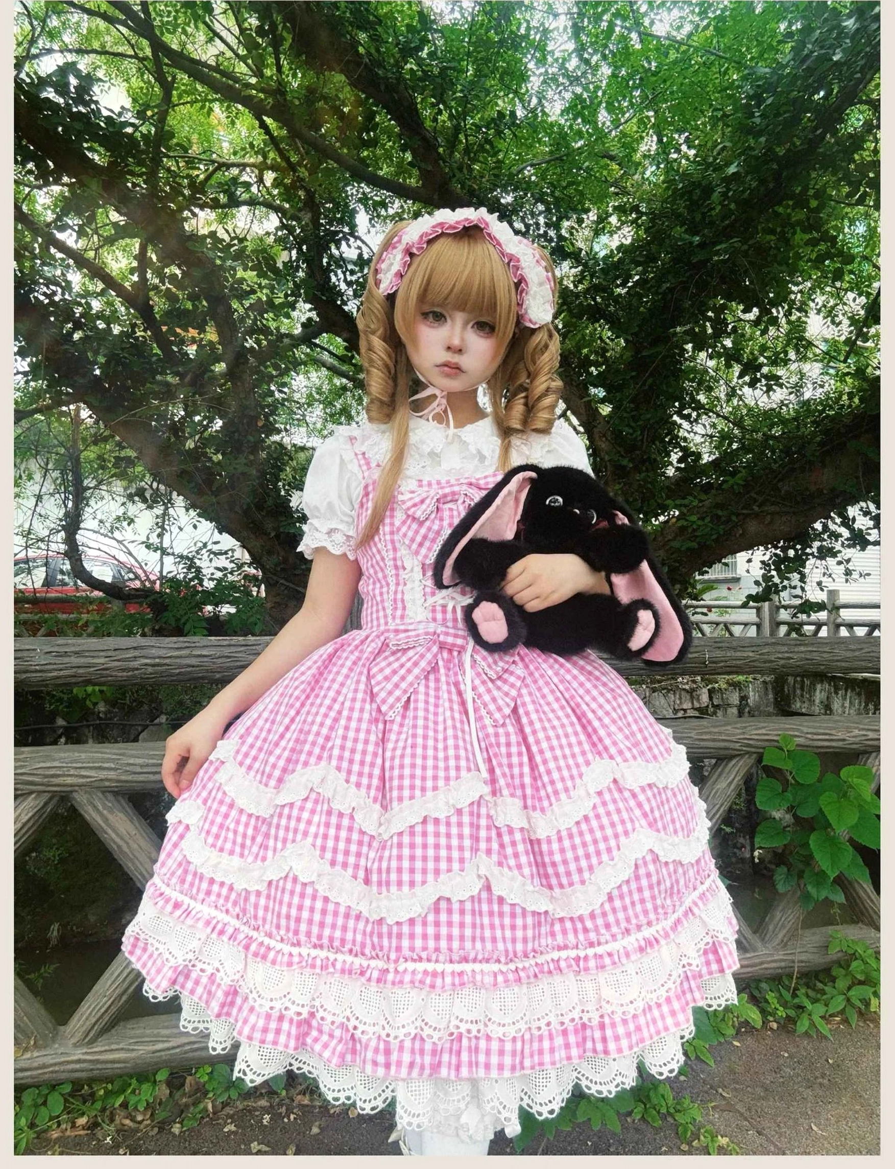Romantic old school Lolita jumper skirt in pink gingham with detailed lace trim and structured corset bodice, full-body side view outdoor garden setting with matching hair clip and black rabbit plush doll