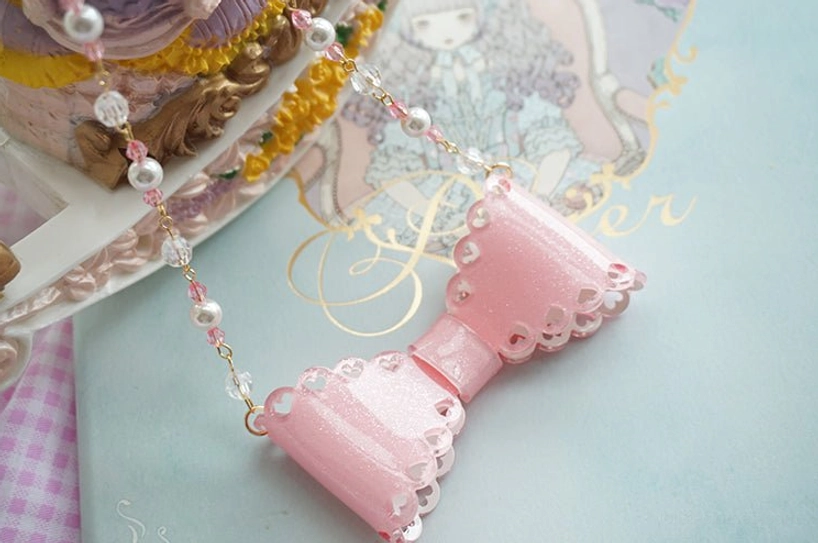 Close-up handmade pink glittery beaded bow necklace with faux pearls and crystal beads on pastel background with cake and illustrated book