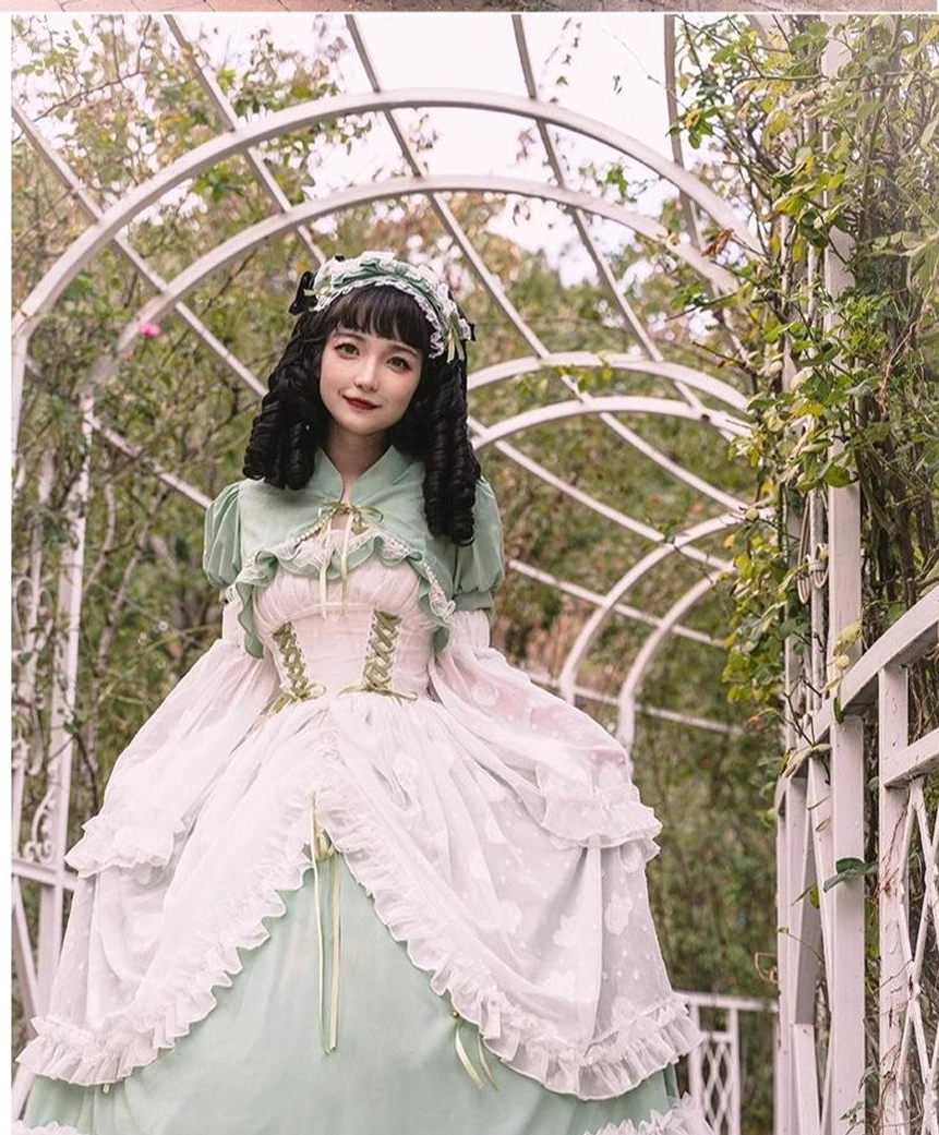 Front full-body view of pink-green bridal lolita dress with white ruffled capelet, pink and white color scheme, corset bodice, garden arch background, and layered skirt