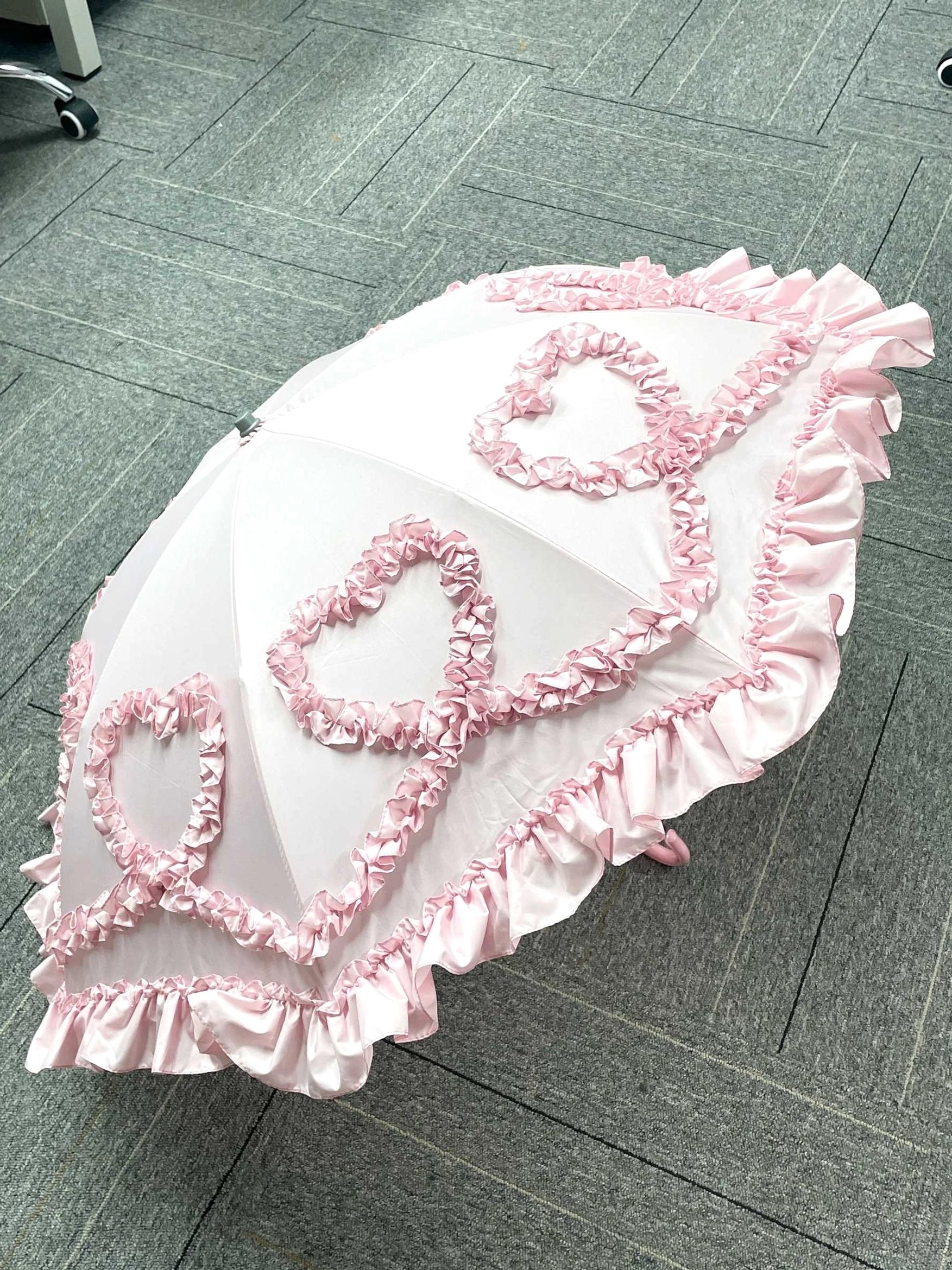 Pink heart-shaped ruffled Lolita parasol with 3D frill embellishments and pompom handle tip displayed on indoor tiled floor, overhead flat lay view showcasing delicate Victorian-inspired design