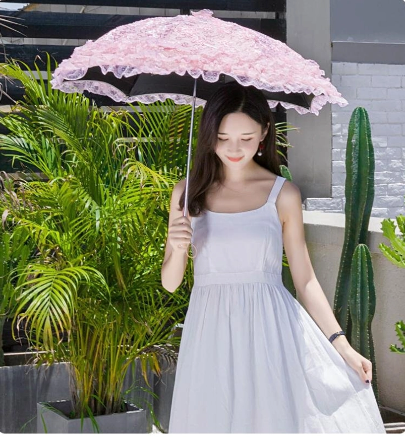 Woman in white slip dress holding pink lace Lolita sun parasol over her head, front eye-level view on outdoor tropical balcony with palm plants and cactus
