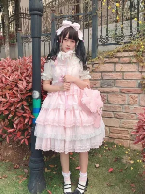 Full-body front view of pink tiered lace lolita dress paired with matching sakura cherry blossom crossbody bag outdoor garden setting