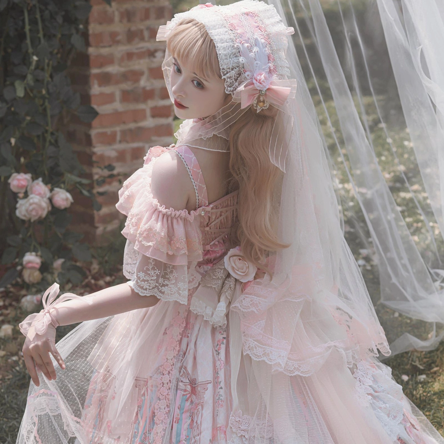 Pink wedding Lolita JSK dress back view with single ruffled off-shoulder strap and lace trims, blonde model with bridal veil and lace bonnet outdoor brick garden