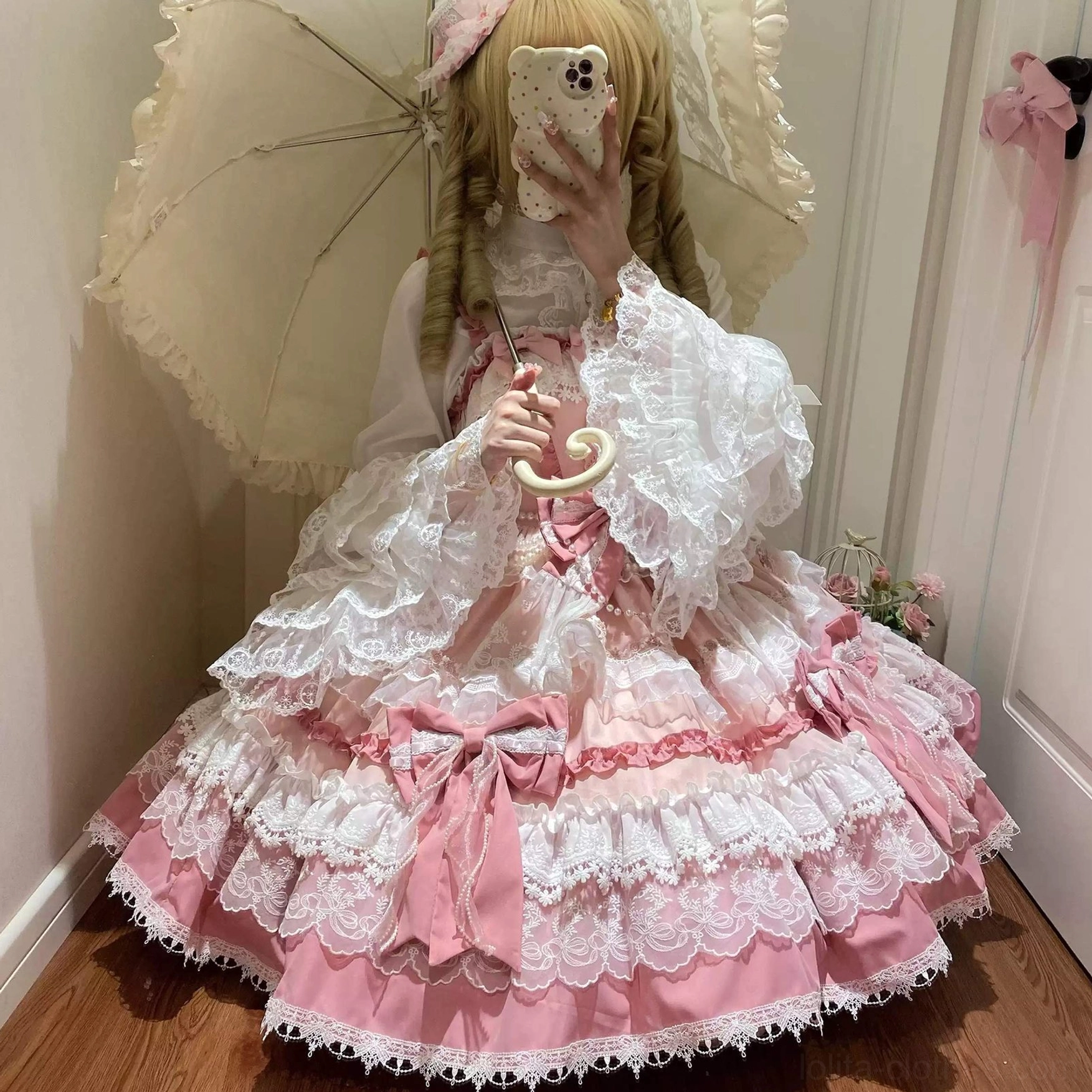Front close-up of pink lolita JSK dress on blonde model holding vintage lace umbrella showing full multi-layered lace skirt, front waist bow, and puff lace sleeves