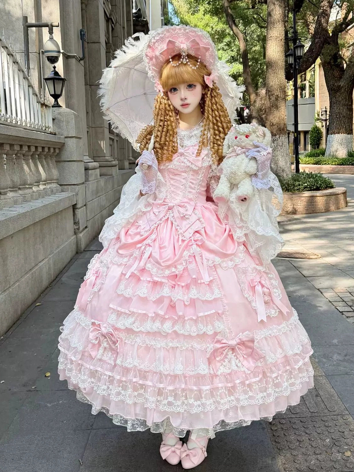Pink sweet wedding Lolita JSK dress overhead front view showcasing lace-trimmed multi-tiered skirt and satin bodice, worn with pink ballet-style shoes and pink bonnet by blonde curly twin tails model in outdoor retro street setting