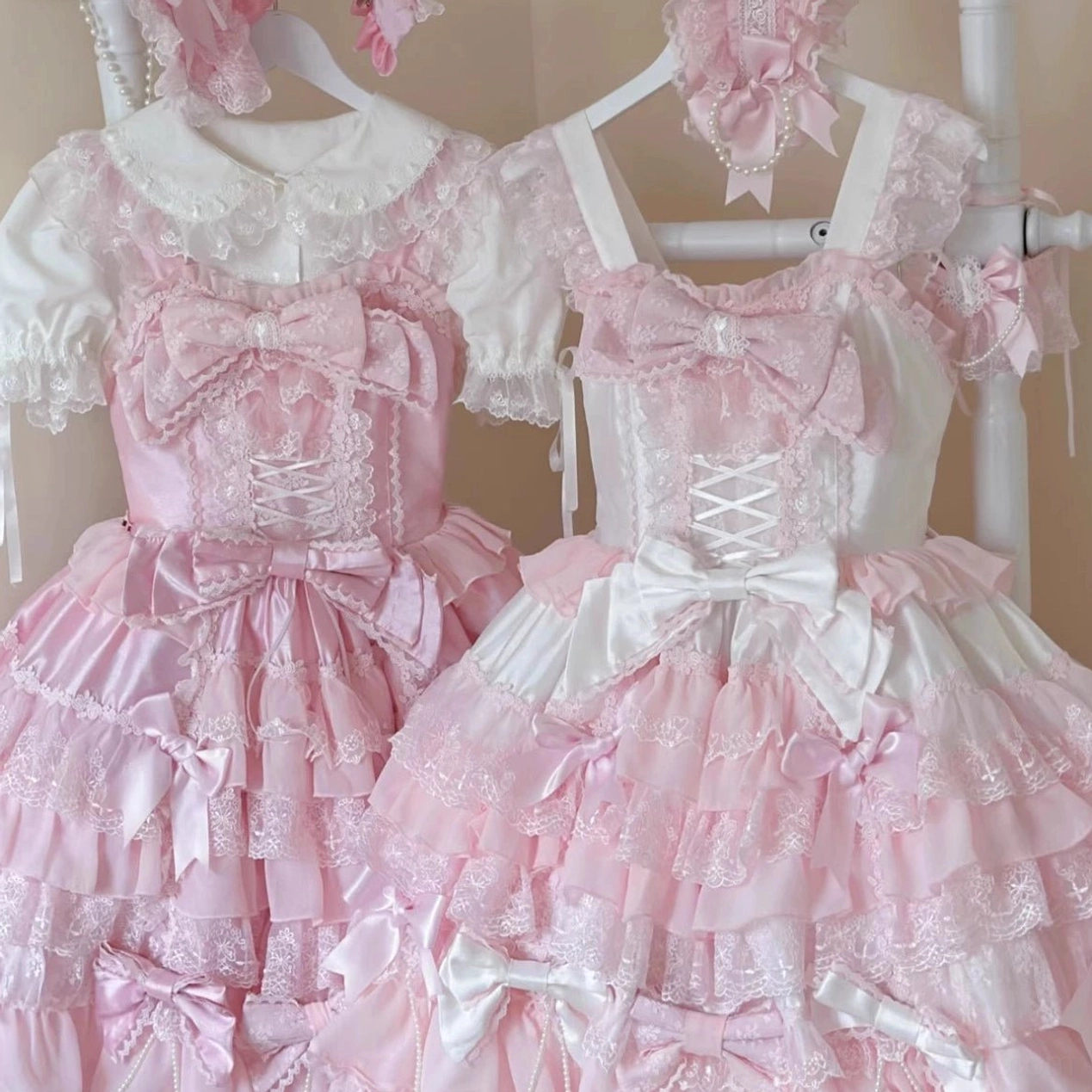 Close-up front hanging view of pink lolita jumper dresses paired with white doll collar blouses showing corset lace-up bodice and multiple decorative satin bows