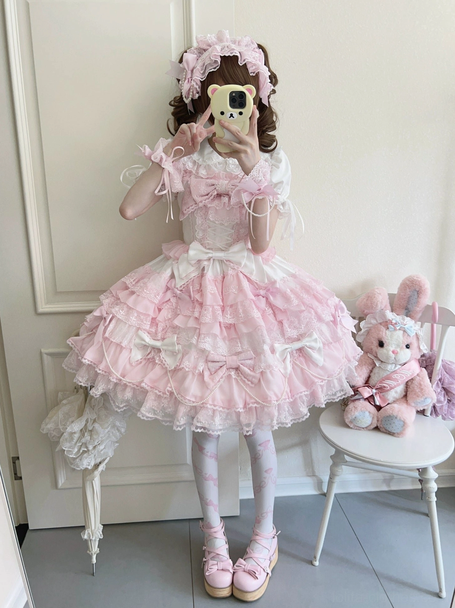 Female model wearing pink lolita dress full-length mirror selfie showing ruffled sleeves sheer patterned tights pink platform loafers home doorway setting