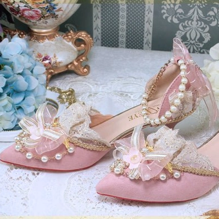 Pink pointed toe Lolita wedding heels overhead flat lay on vintage lace tablecloth with hydrangea flowers and decorative vase, showing pink suede upper, pink flower bow decor, lace ruffles, pearl and gold trim details