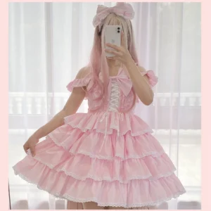 Pink off-shoulder sweet lolita jumper skirt with ruffled sleeves and lace-trimmed tiered cake-style skirt, half-body front view with model in indoor window scene