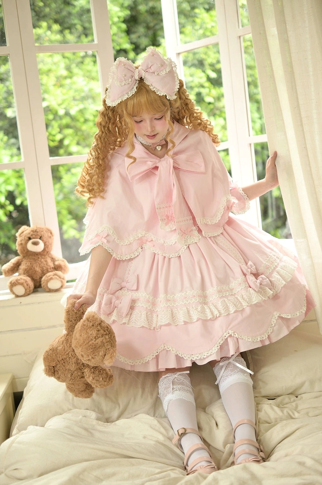 Pink lolita cape dress set front three-quarter shot blonde model standing on bed teddy bear bedroom flared lace-trimmed sleeves ruffled cape collar large front bow