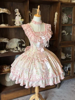 Front left three-quarter view of pink rose print classic Lolita JSK dress with ruffled square neckline, front satin bows, and tiered ruffled skirt hem displayed on vintage wooden curio cabinet