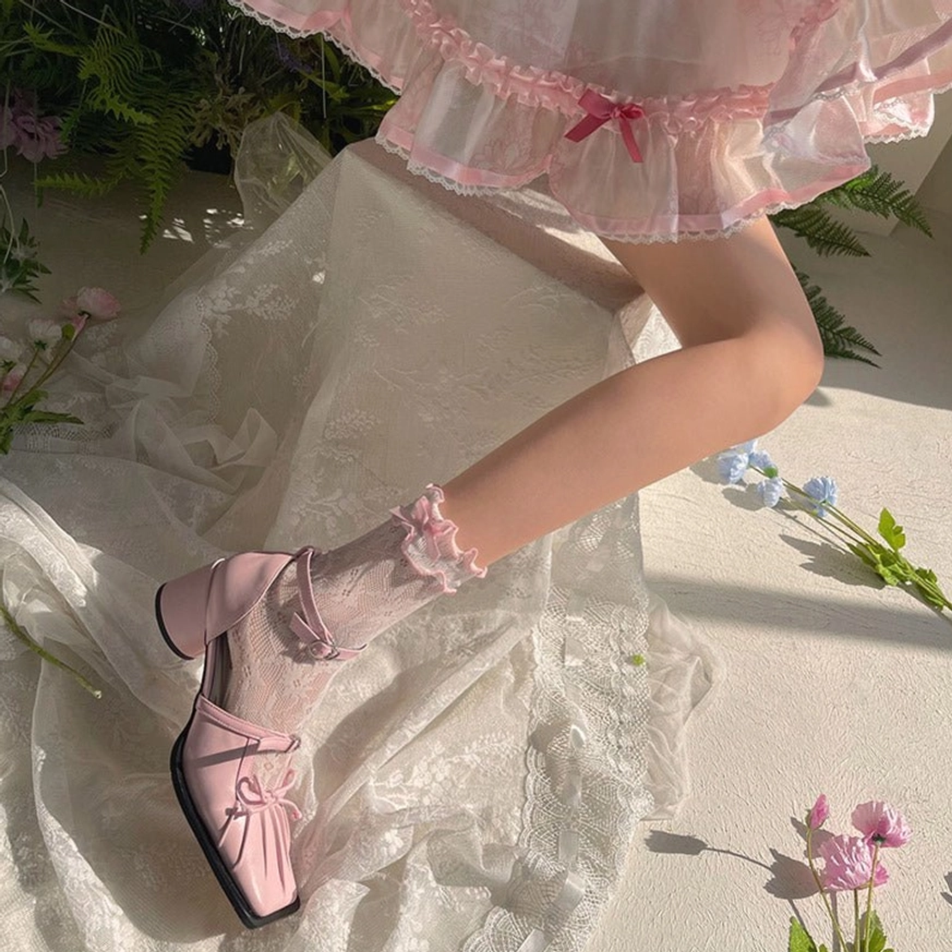 Light pink ruffled lace ankle socks with delicate floral pattern worn with pink frilly Lolita skirt on model's leg side view with sheer fabric and wild flowers backdrop