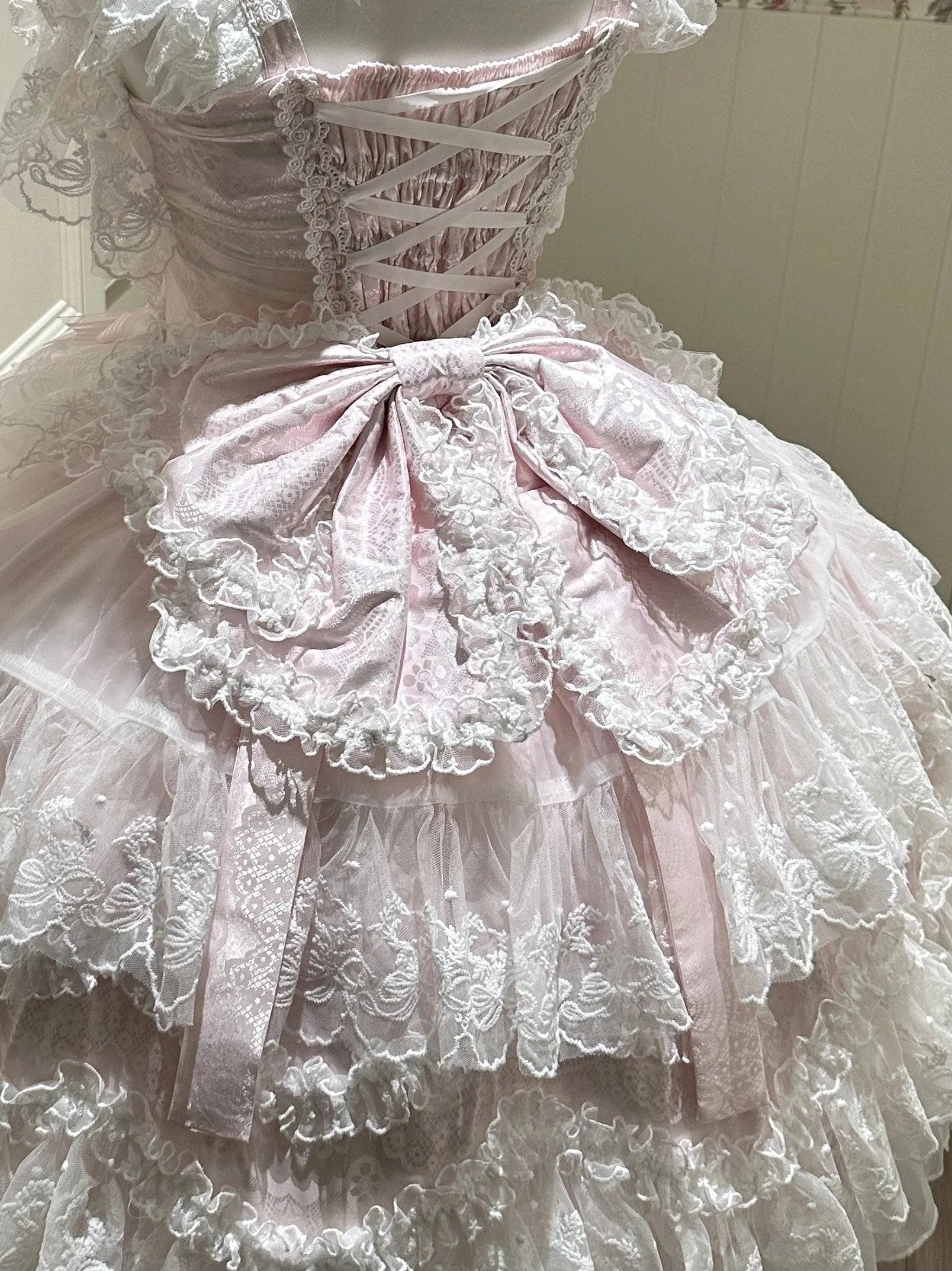 Back detail close-up view of pink Sakura Blossom Dream Sweet Lolita bow trailing accessory showing oversized layered lace back bow, corset-style lace-up waist design, draped satin ribbons, and voluminous ruffled tulle skirt layers with coordinating lace trim for dramatic Sweet Lolita silhouette
