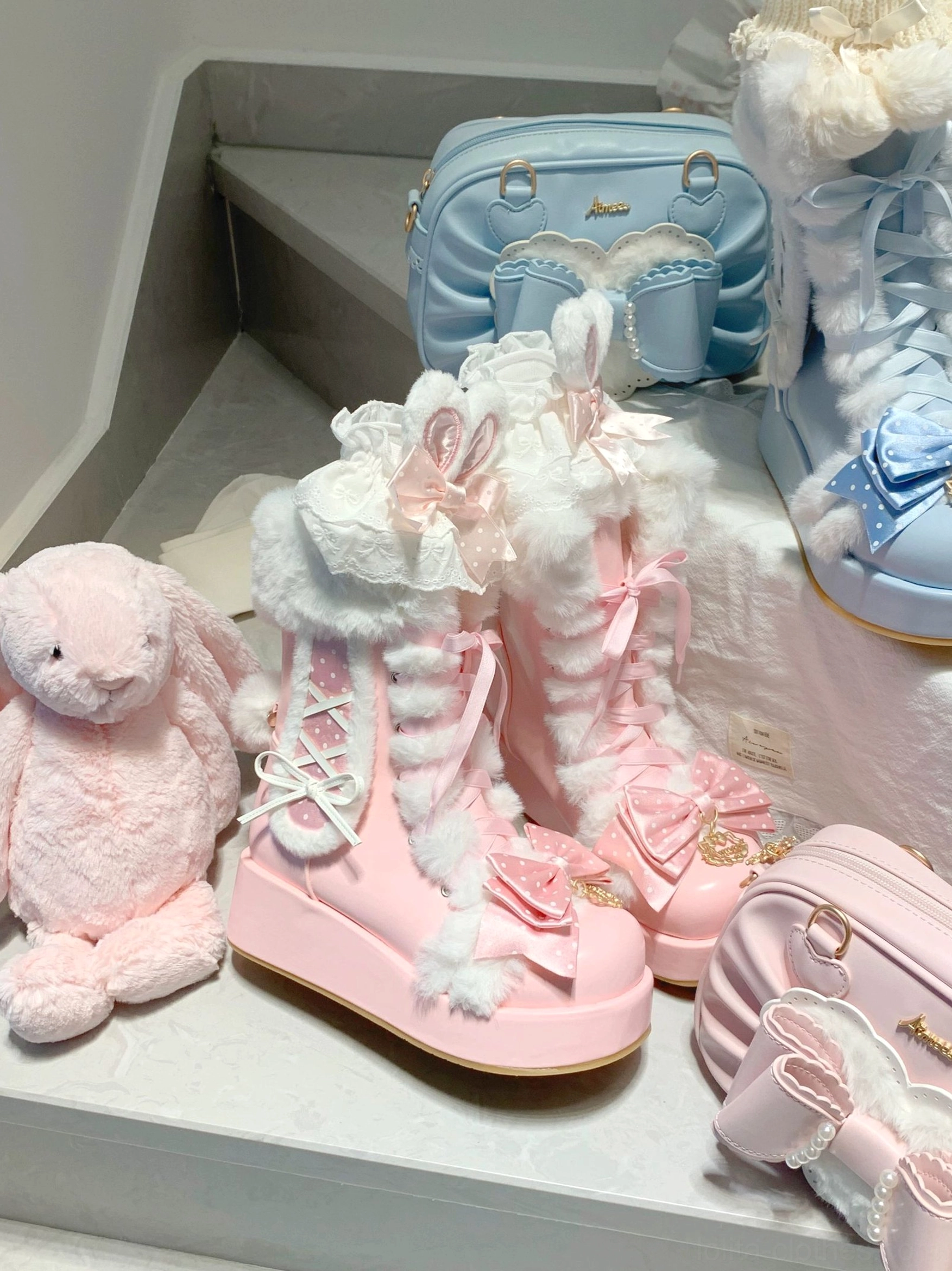 Overhead flat lay of pink and sky-blue sweet lolita platform boots with matching bags and plush rabbit decor on home staircase, showing furry collar, lace-up front, bow decorations, and elevated platform soles