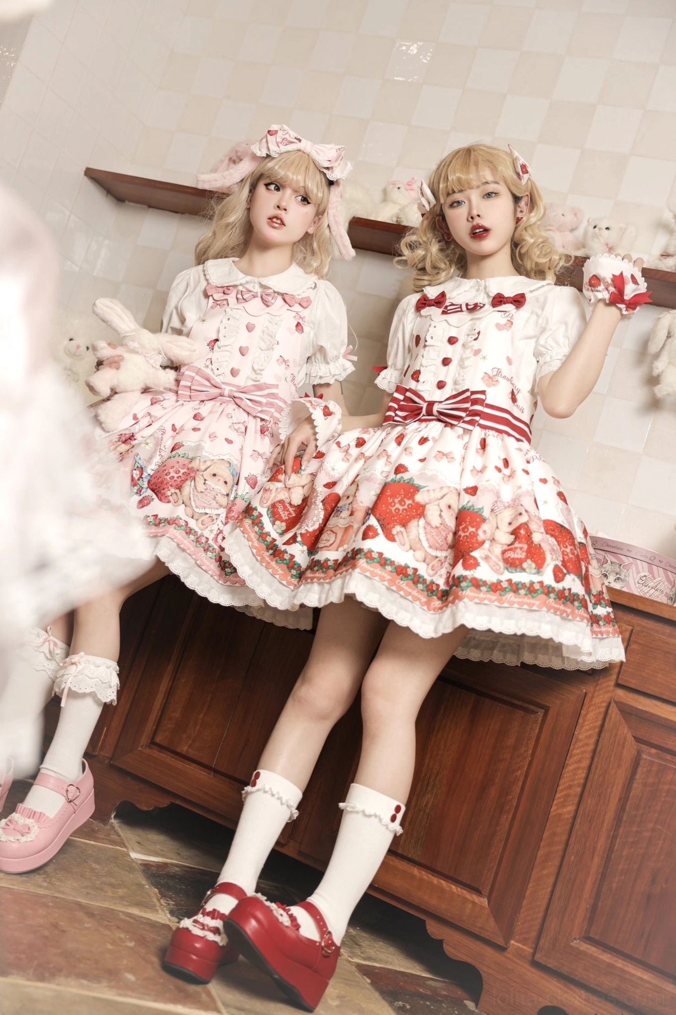 Two blonde models wearing matching pink strawberry rabbit lolita dresses with puff sleeves red bow waistbands and ruffled skirt hems leaning on wooden cabinet