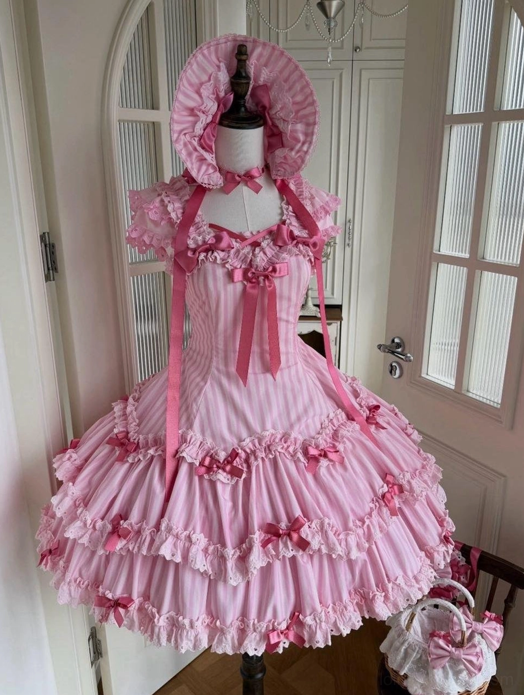 Pink vertical striped classic old school Lolita JSK dress front view on mannequin with ruffled skirt, bow accents, and matching bonnet and handbag accessories