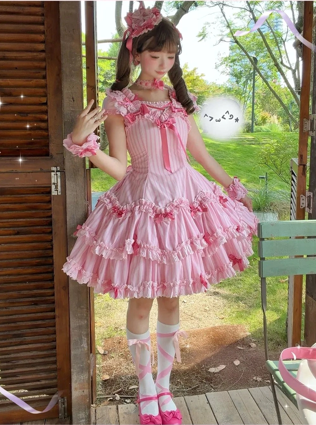 Front full-body outdoor photo of pink striped old school Lolita dress beside wooden shutter door with twin tails hairstyle and ruffled skirt bow details