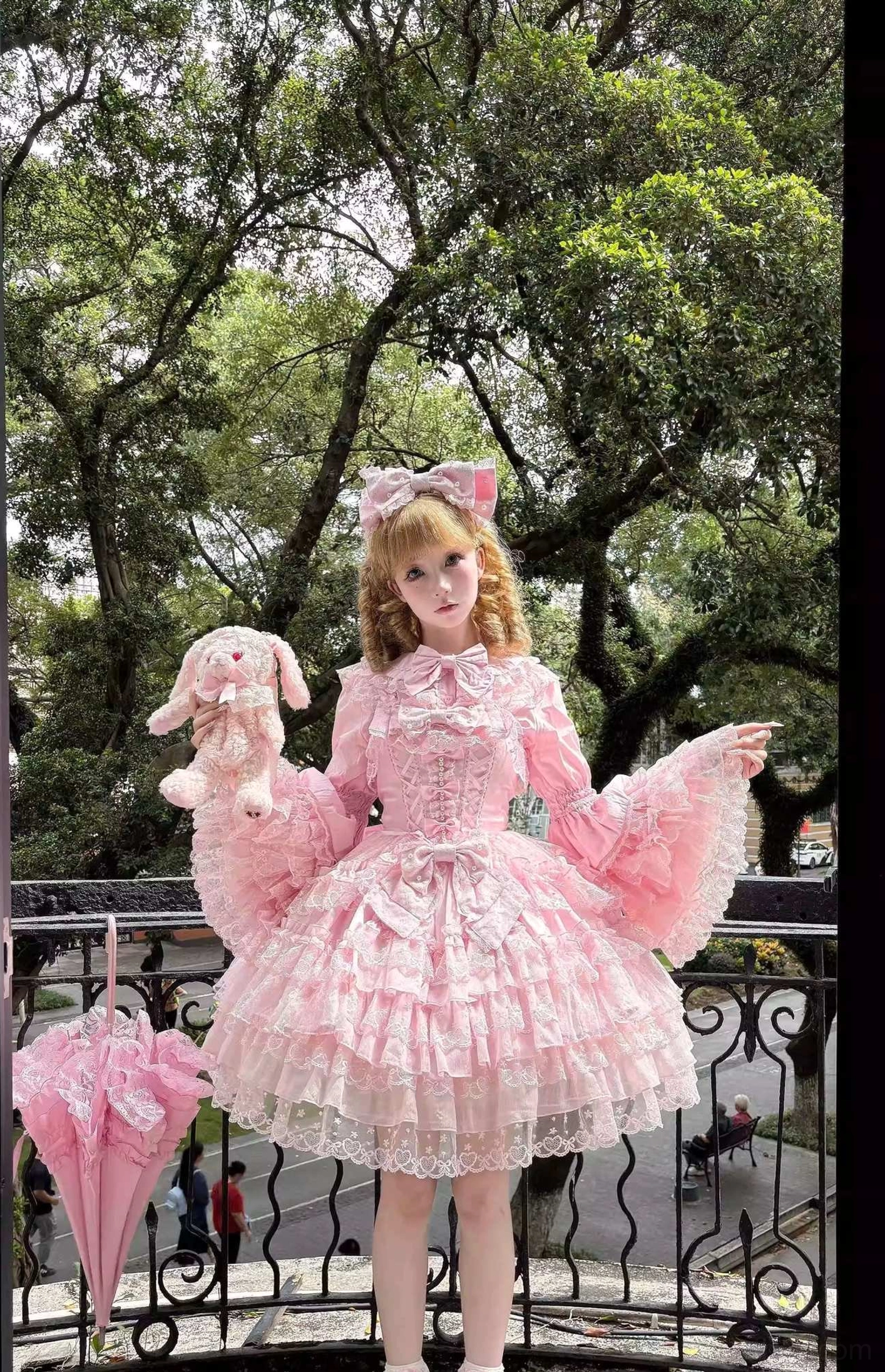 Pink Sweet Lolita dress accessory set full front standing on outdoor balcony with tiered ruffle lace skirt, frilled flared sleeves, matching hair bow, foldable lace parasol, and plush rabbit