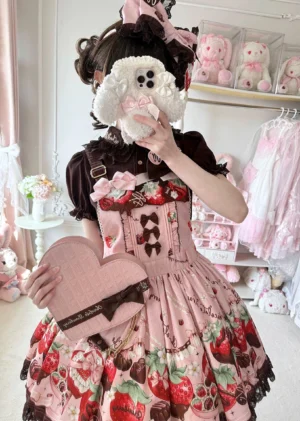 Pink sweet Lolita heart-shaped handbag worn by model in strawberry print dress with matching hair accessories in pastel bedroom setting with lace decorations