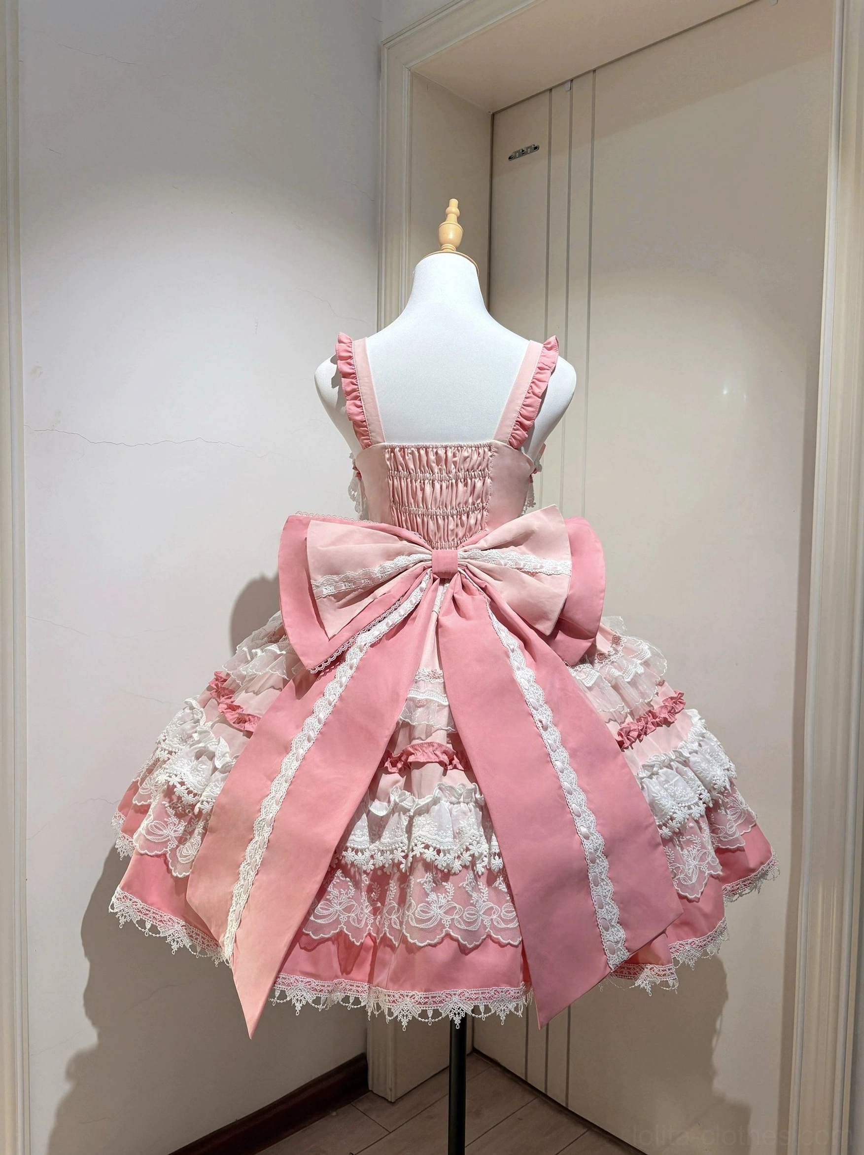 Back view of pink sweet lolita JSK dress on mannequin displaying large dramatic trailing oversized bow, layered lace trims, and adjustable spaghetti shoulder straps