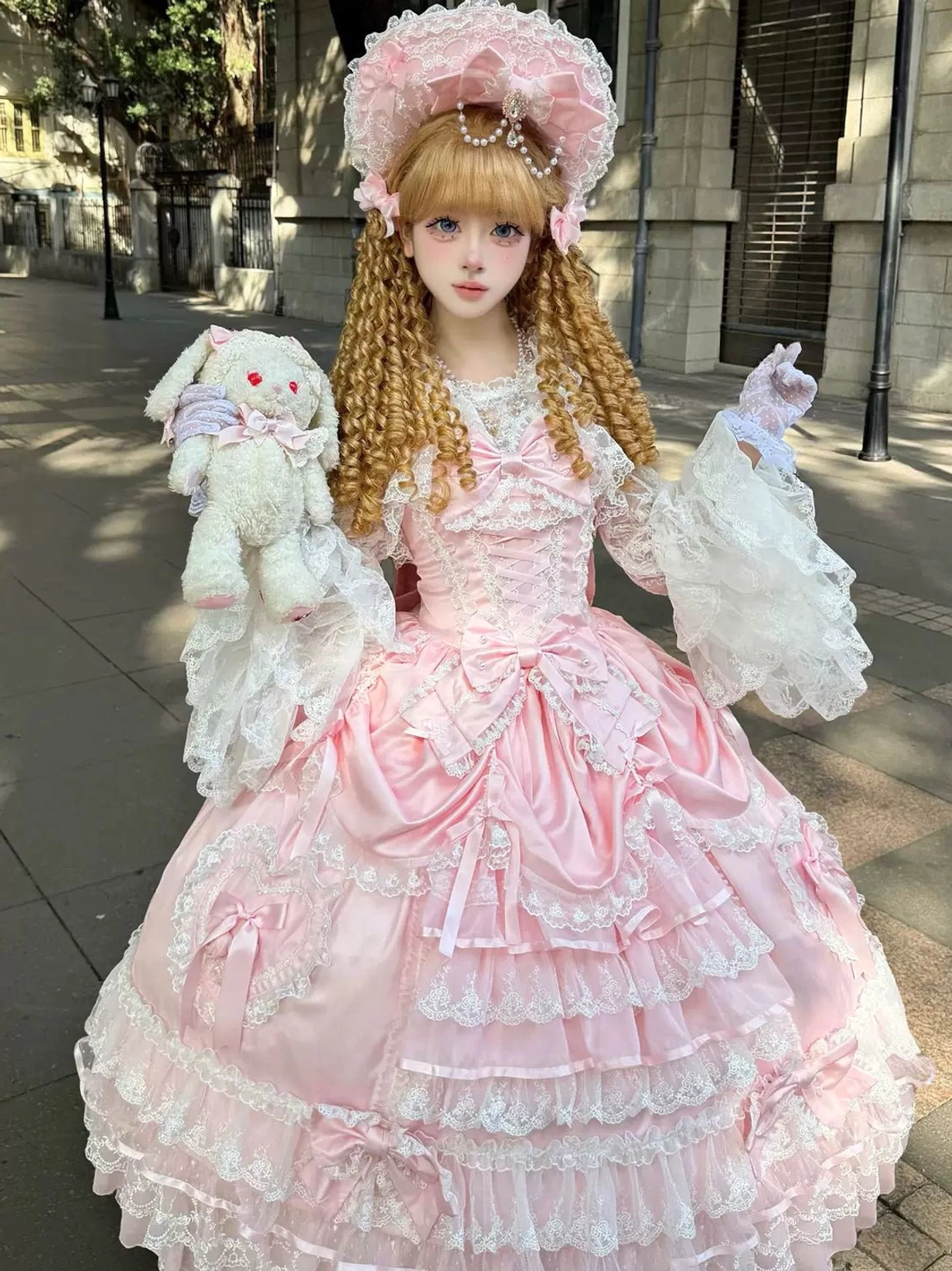 Pink sweet wedding Lolita JSK dress full-body front view with tiered lace ruffle skirt, sweetheart bodice bow details, detachable lace arm sleeves, worn by blonde model with pink bonnet holding plush rabbit and lace gloves