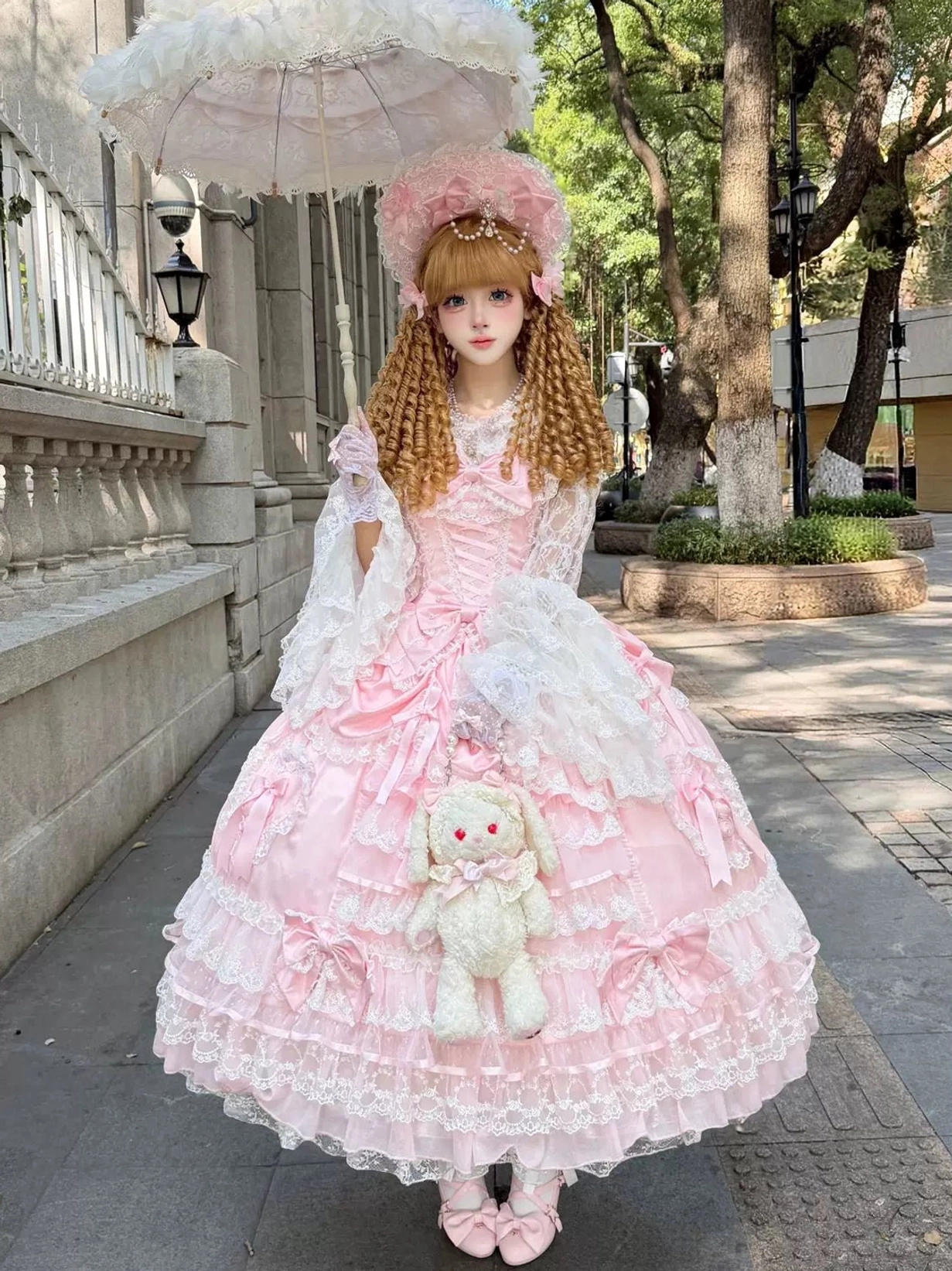 Pink sweet wedding Lolita JSK dress with tiered lace skirt, waist bow decorations, worn with pink bonnet, matching pink footwear, lace gloves, and carrying lace parasol and plush rabbit handbag in outdoor street setting