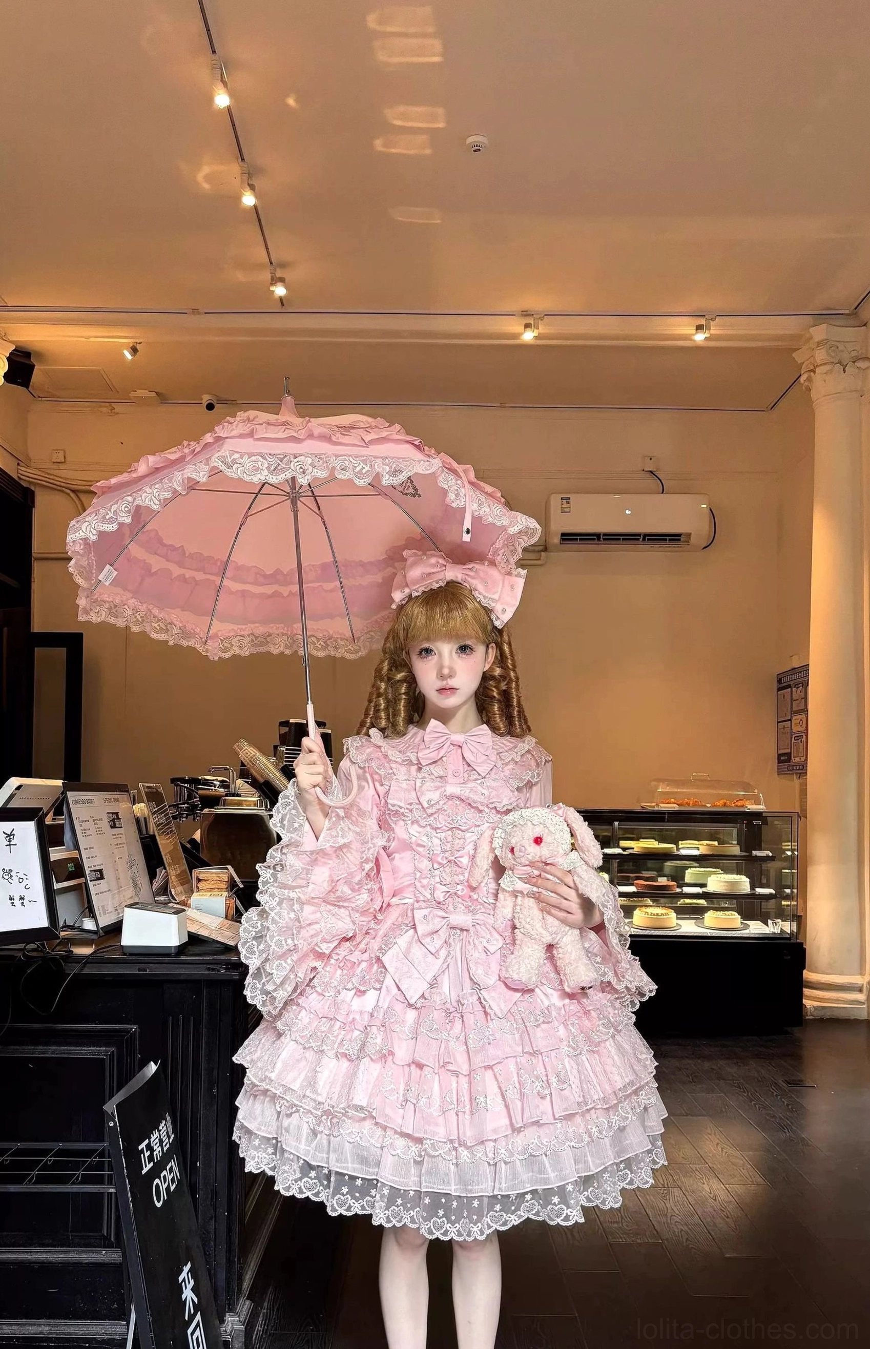 Pink Sweet Lolita JSK dress full-body front view with tiered ruffle lace skirt, matching hair bow, and frilled inner blouse worn in bakery setting with parasol and plush rabbit accessory