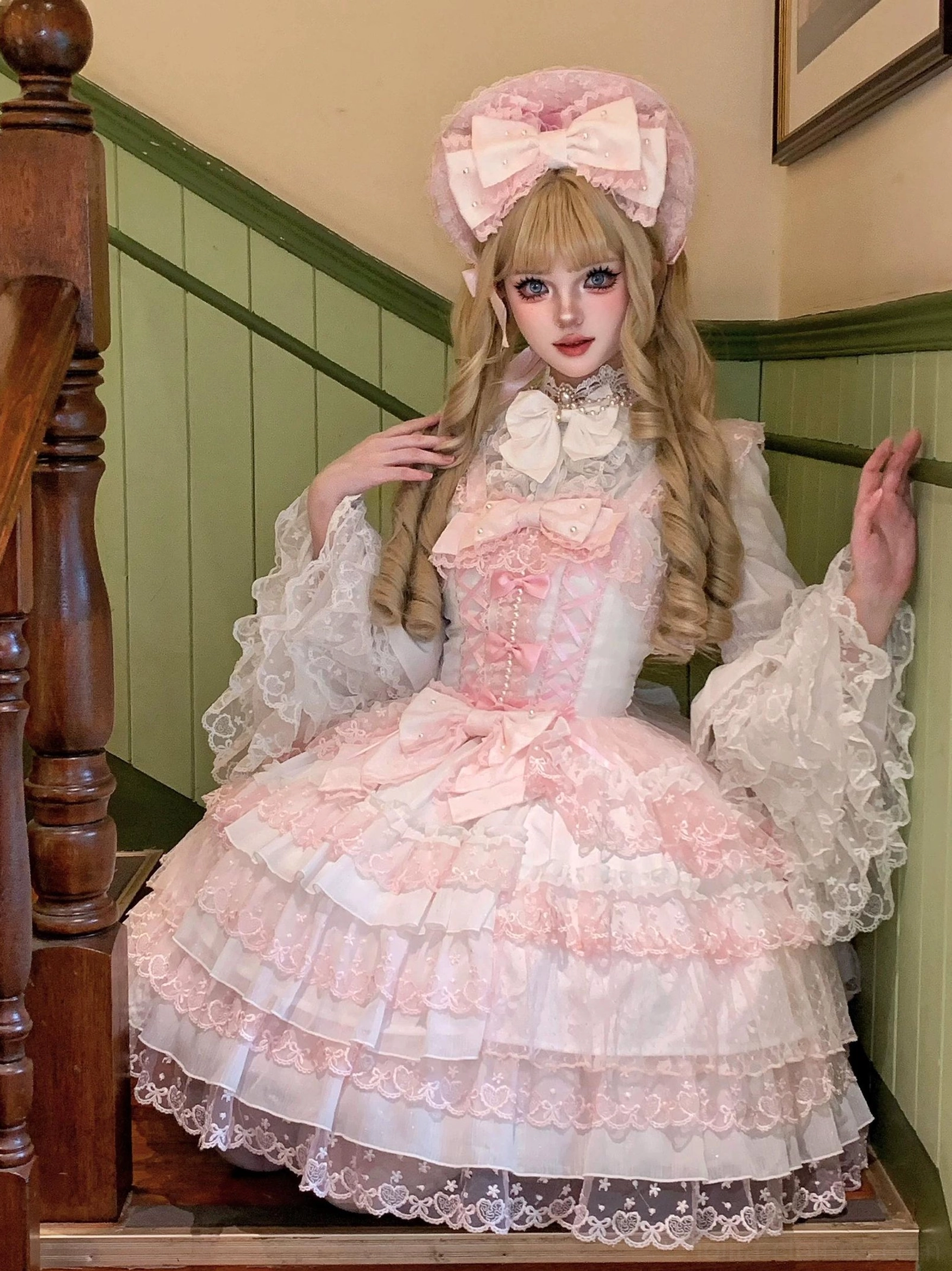 Pink Sweet Lolita formal JSK dress set standing front view on staircase with tiered layered lace skirt, large front bows, frilled flared lace sleeves, and big hair bow
