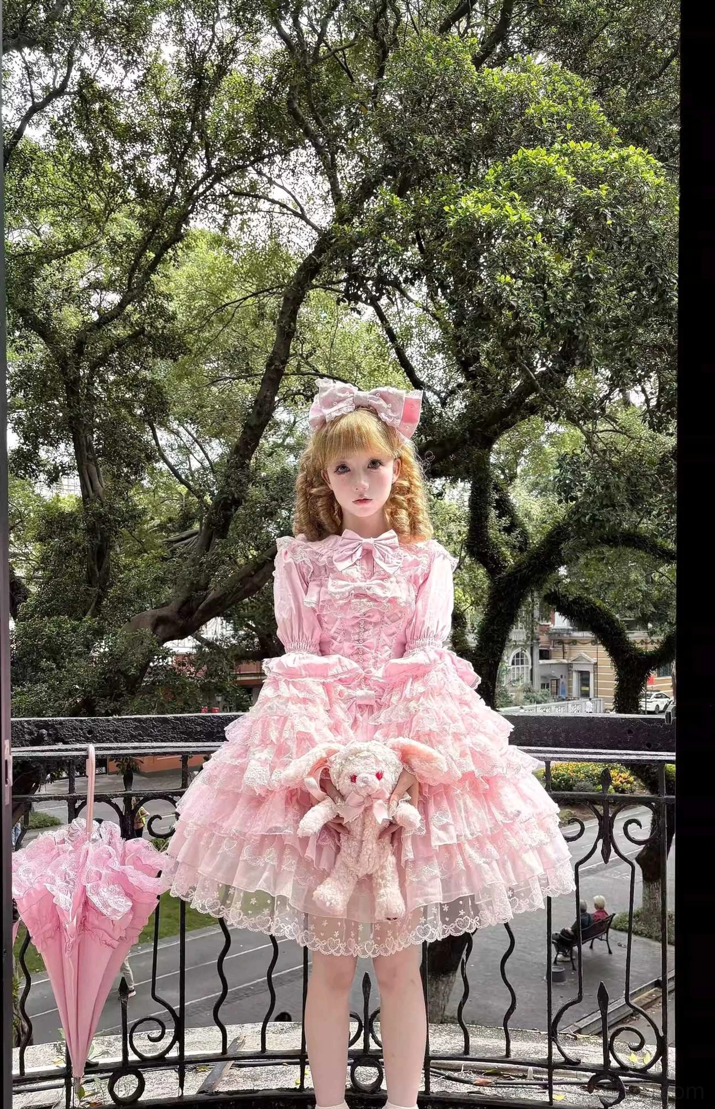 Pink Sweet Lolita JSK dress set full front standing view on outdoor balcony with tiered lace hem, front waist bow, attached plush rabbit decor, and foldable lace parasol